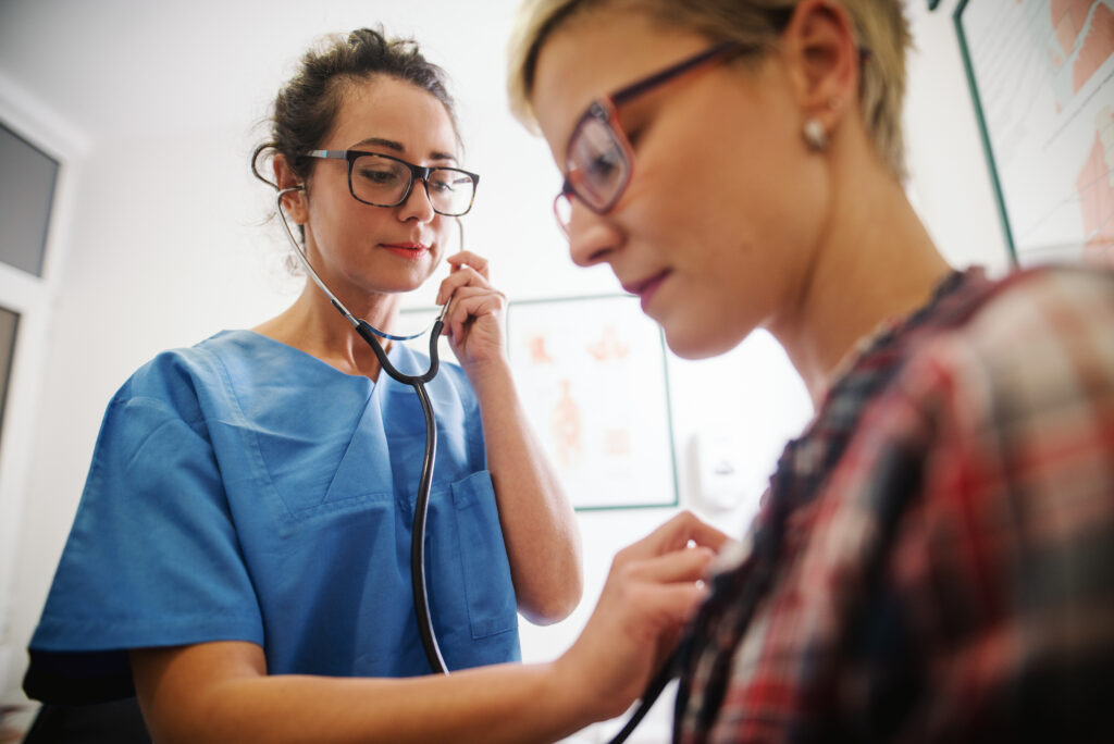A female doctor or nurse in blue scrubs and glasses listens to a female patient's heart or lungs with a stethoscope in an examination room.