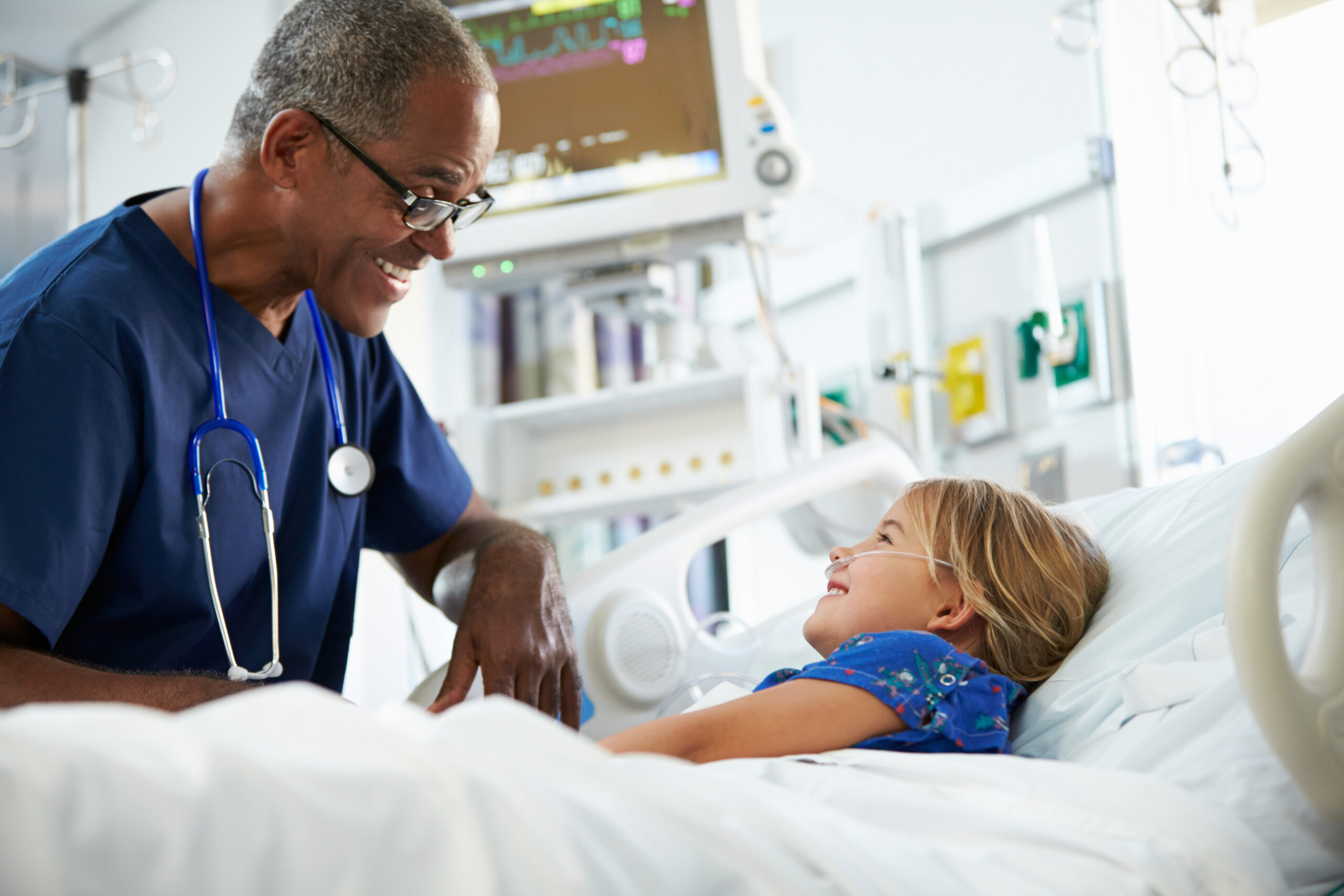A smiling, kind male doctor leans over a hospital bed to speak with a happy young girl patient with a nasal cannula. A vital signs monitor is visible in the background.