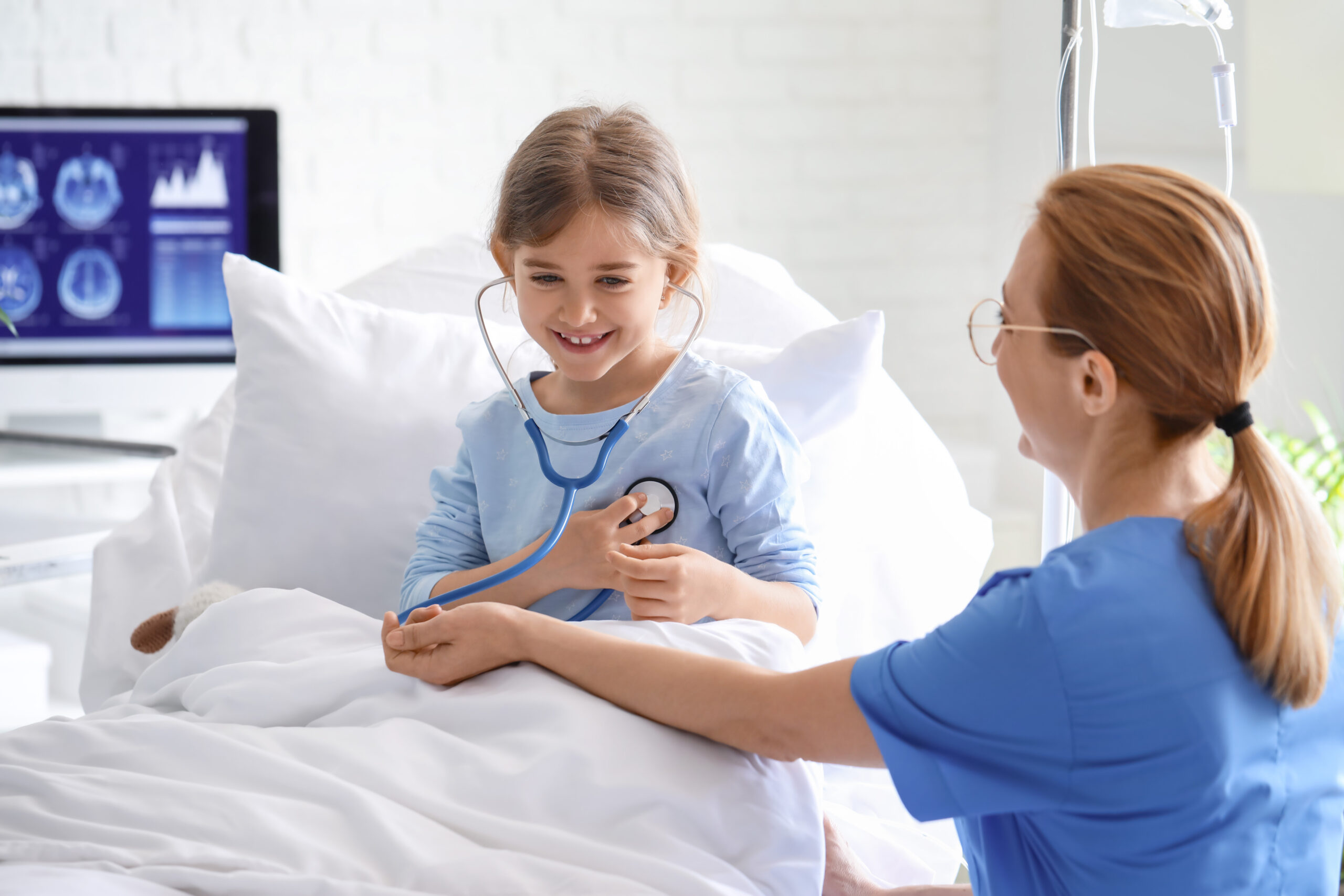 A happy young girl sitting up in a hospital bed is playing doctor by using a stethoscope to listen to the arm of a smiling female nurse.