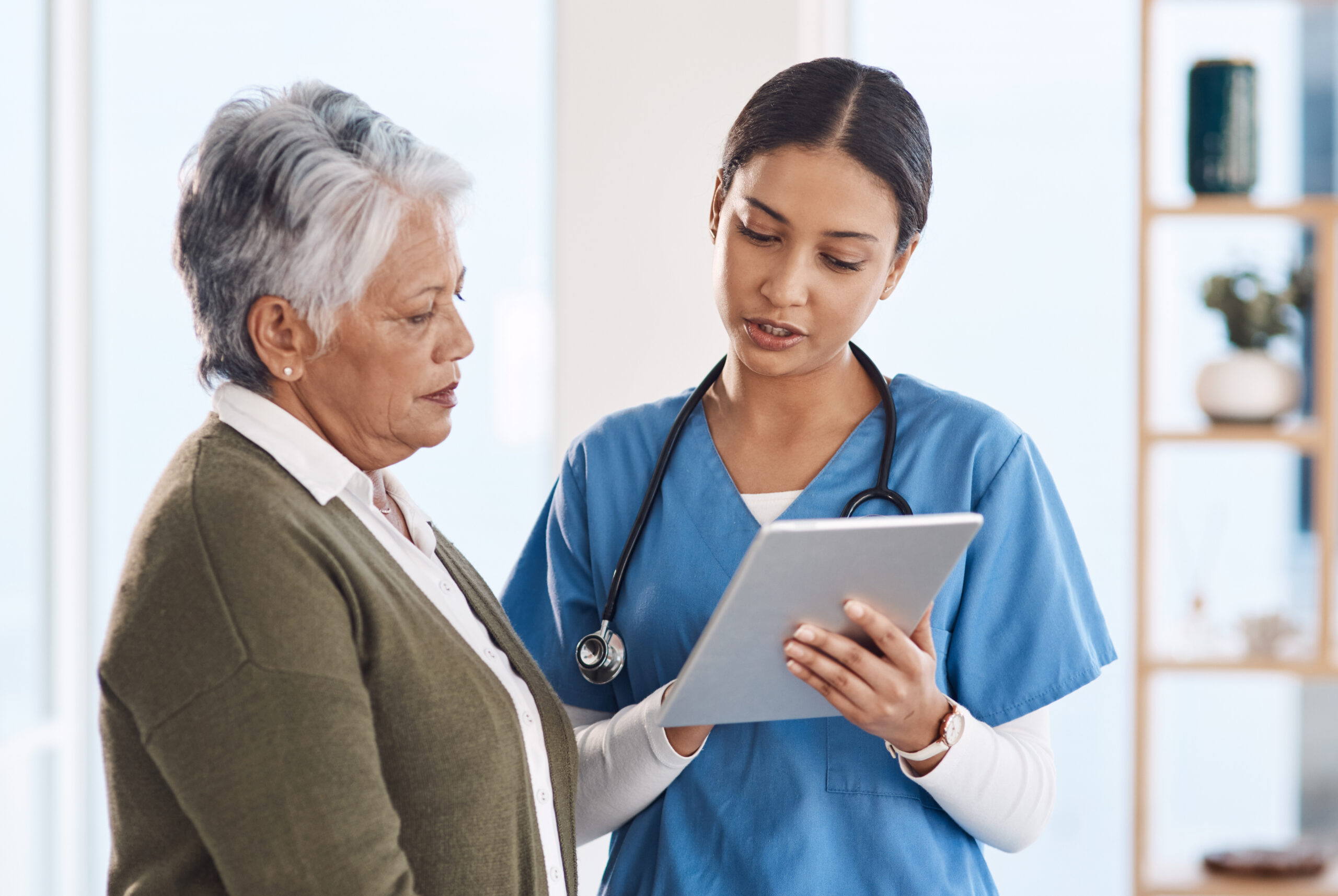 A young female doctor in blue scrubs and a stethoscope is showing information on a tablet to an older female patient with gray hair.