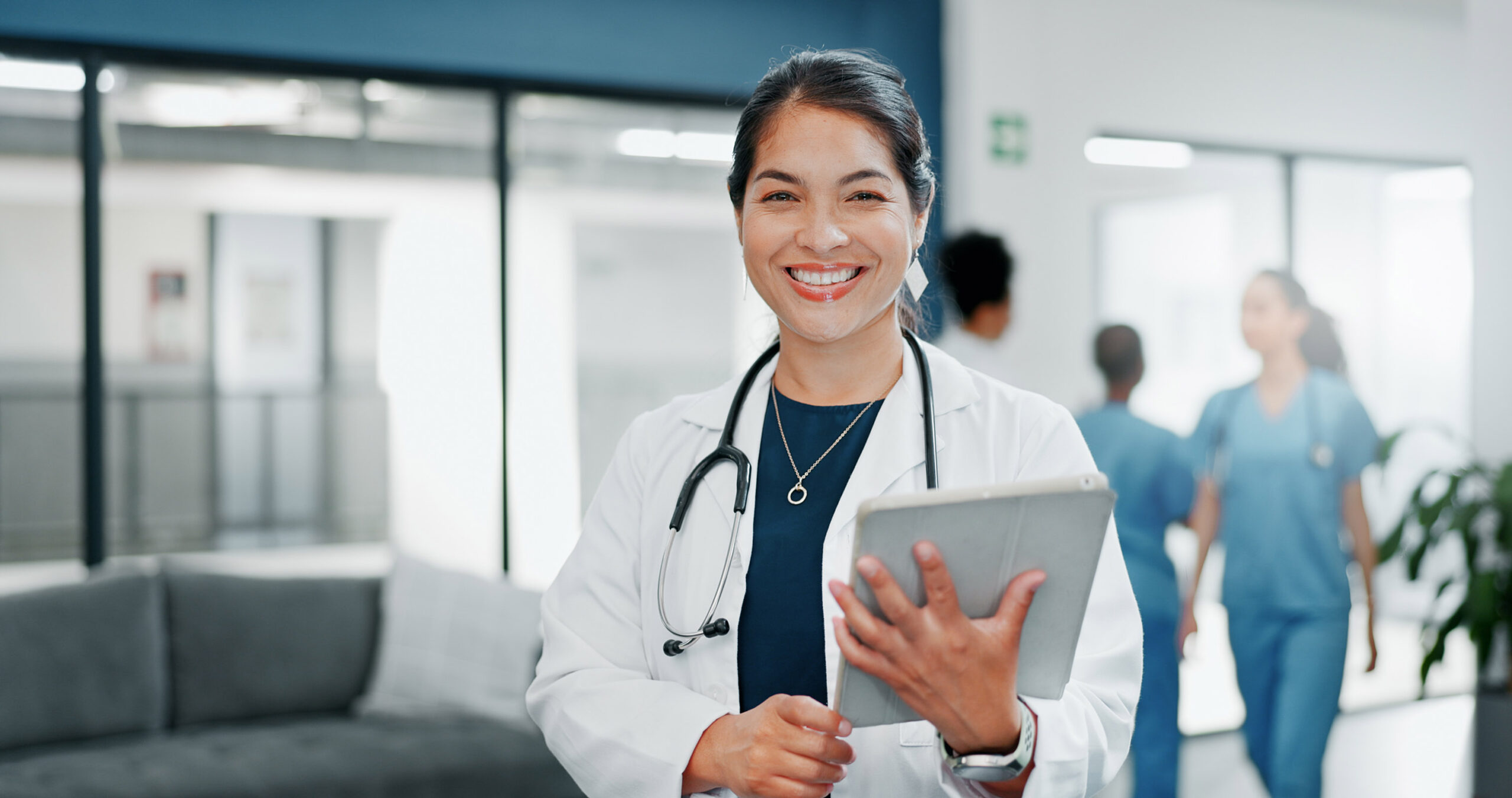 A smiling doctor wearing a white coat and stethoscope holds a tablet in a modern hospital setting, with other healthcare professionals in blue scrubs walking in the background.