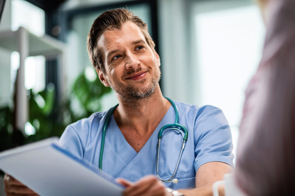 A smiling male doctor in blue scrubs with a stethoscope around his neck holds a clipboard and talks to a patient in a bright, modern office.