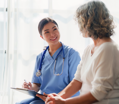 A nurse in blue scrubs with a stethoscope smiles while holding a clipboard and talking to an older woman in a white sweater. They are sitting indoors in a well-lit room.
