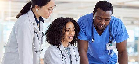 A team of three diverse medical professionals—two doctors in white lab coats and one nurse in blue scrubs—collaborate and look down at a project or chart.
