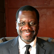 A man wearing a dark suit, white shirt, and black tie smiles while seated indoors against a reddish-brown wall.