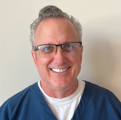Smiling middle-aged man with short gray hair and glasses, wearing blue scrubs, standing in front of a plain light-colored wall.