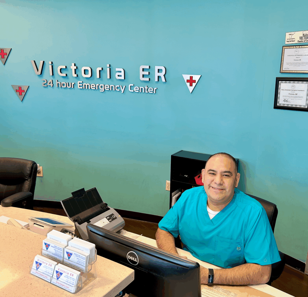 A young woman in a red 'Victoria ER' shirt smiles at a male patient or visitor checking in at a reception desk.