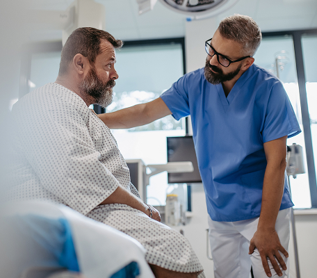 A doctor in blue scrubs gently talks to a concerned male patient sitting on a hospital bed in a medical exam room. The doctor has a reassuring hand on the patient’s shoulder.