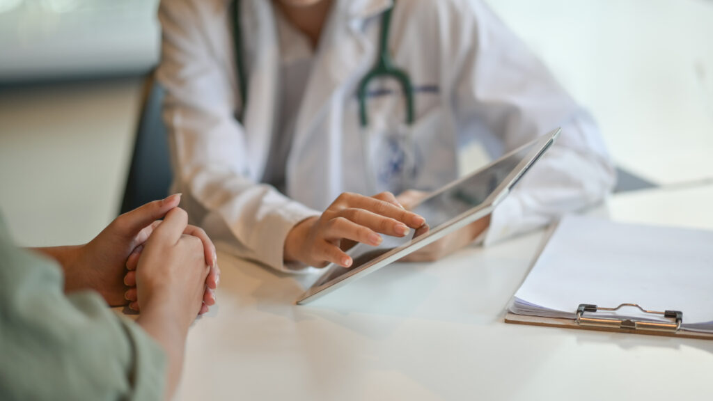A doctor in a white coat, holding a tablet, discusses information with a patient. Both are seated at a desk, with a clipboard and papers nearby, suggesting a medical consultation.