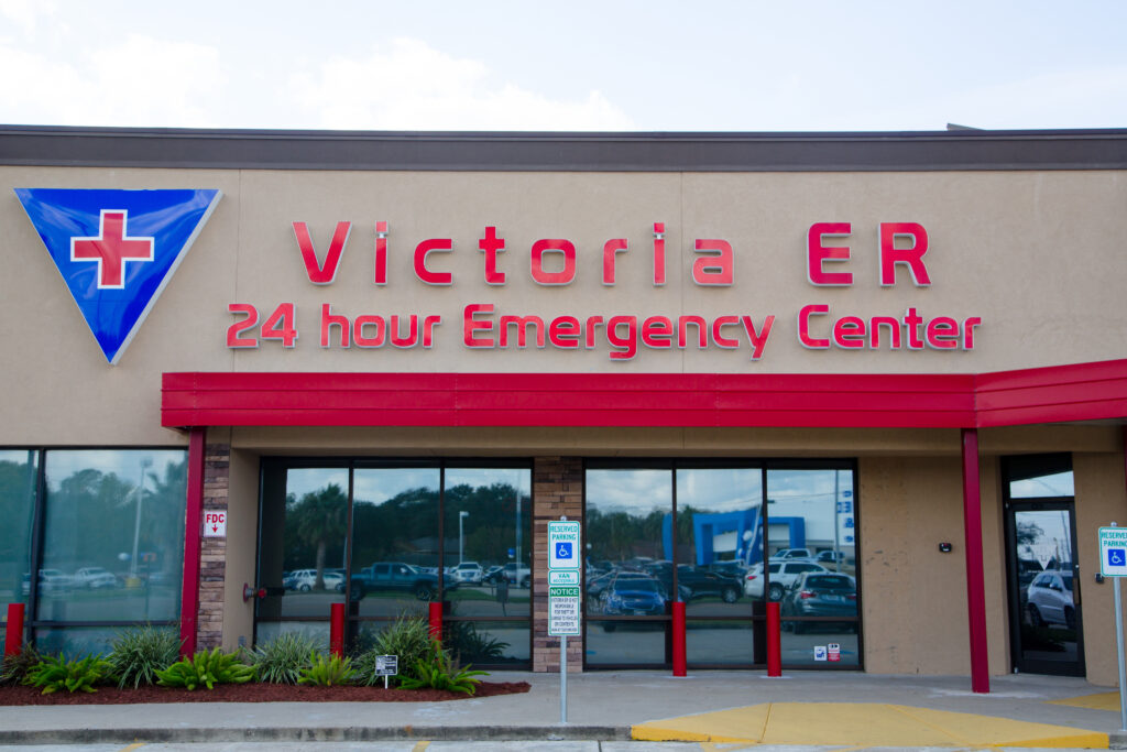 The exterior sign for 'Victoria ER 24 hour Emergency Center' with a blue and red medical cross logo next to the name, and a reserved parking sign for people with disabilities visible near the entrance.