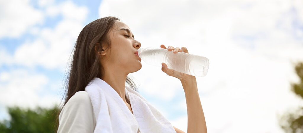 A woman with a white towel around her neck drinks water from a clear plastic bottle outdoors, with a bright sky and green trees in the background.