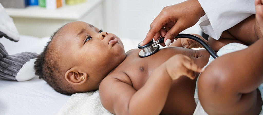 A doctor uses a stethoscope to listen to the chest of a baby lying on an exam table. The baby looks up at the doctor while wearing only a diaper.