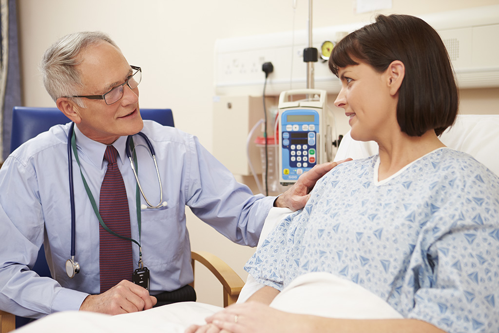 A doctor with gray hair and glasses speaks reassuringly to a female patient in a hospital bed, gently touching her shoulder. The patient wears a hospital gown and smiles back at the doctor in a medical setting.
