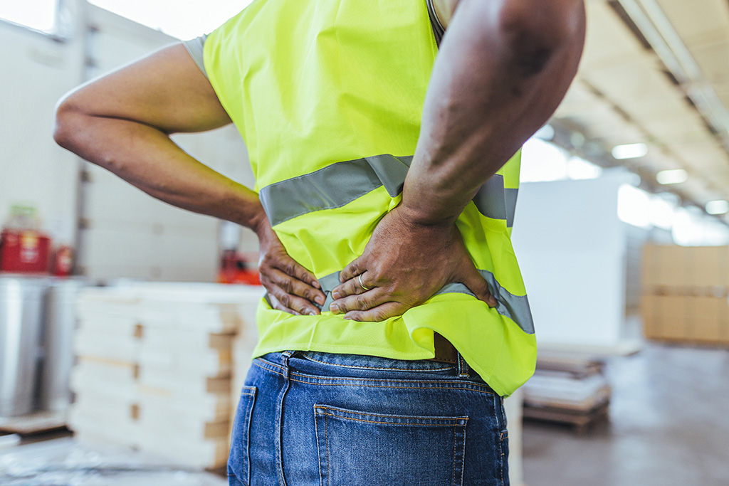 A person wearing a yellow safety vest holds their lower back with both hands, appearing to be in pain, in an industrial or warehouse setting.