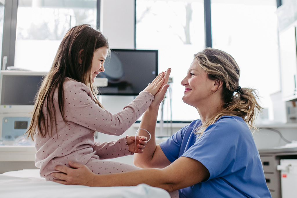 A smiling nurse in blue scrubs gives a high-five to a young girl sitting on a hospital bed. The girl is wearing pink pajamas and both appear happy. Medical equipment is visible in the background.