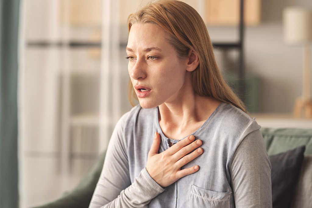 A woman with long blonde hair sits indoors, holding her hand to her chest and appearing concerned or short of breath. She wears a light gray top and looks slightly to the side.
