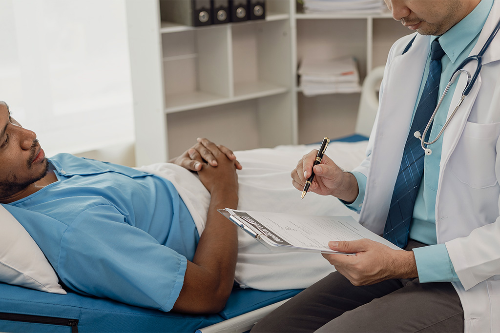 A doctor in a white coat holding a clipboard and pen sits beside a patient lying on a medical bed. The patient is wearing a blue hospital gown with hands folded on the stomach. Shelves are visible in the background.