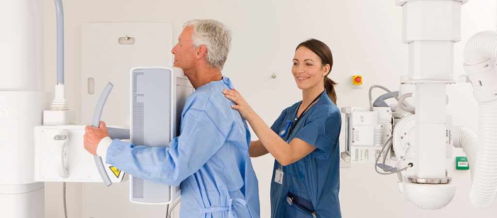 A healthcare worker assists an older man in a hospital gown as he stands for a chest X-ray in a medical imaging room. The worker is smiling and guiding the patient, who faces an imaging machine.