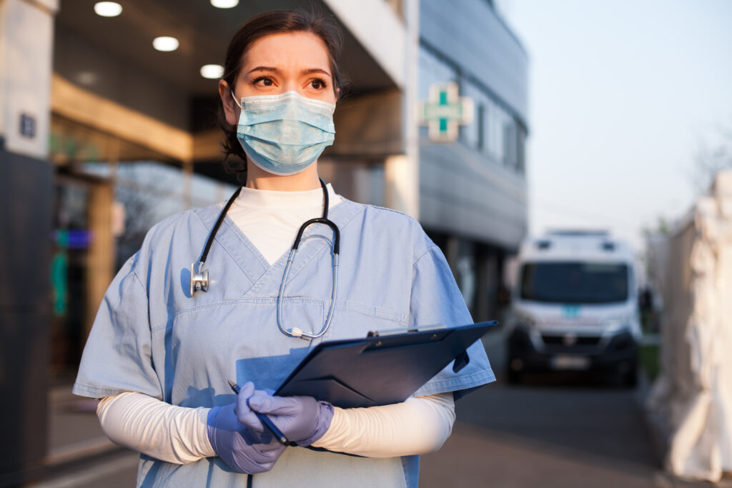 A healthcare worker wearing scrubs, gloves, a mask, and a stethoscope stands outside a medical facility holding a clipboard. An ambulance and a building are visible in the background.