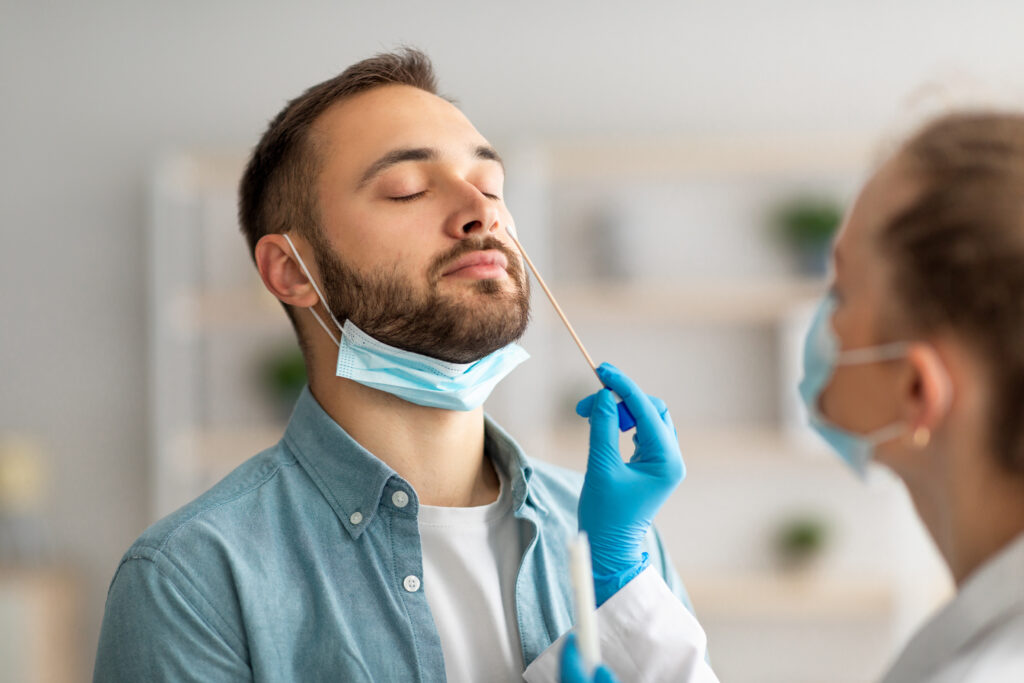A medical professional in blue gloves and a surgical mask is administering a nasal swab test to a man. The man, also wearing a surgical mask pulled down below his nose, has his eyes closed while the professional holds the swab near his nostril. The scene suggests a COVID-19 test or similar diagnostic procedure.