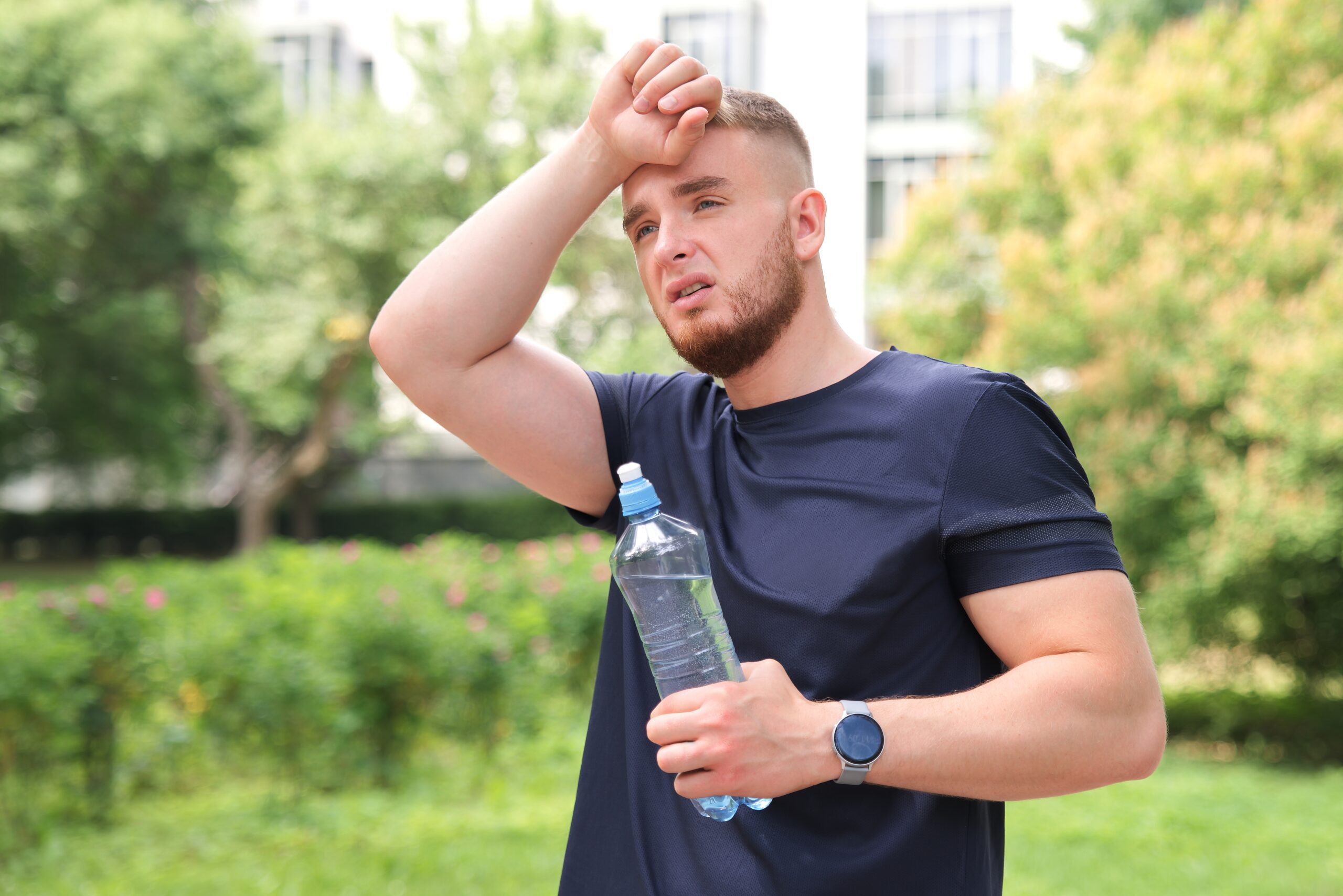 A tired, sweaty man in a dark blue athletic shirt stands outdoors, wiping his forehead with his arm and holding a clear water bottle in his other hand.
