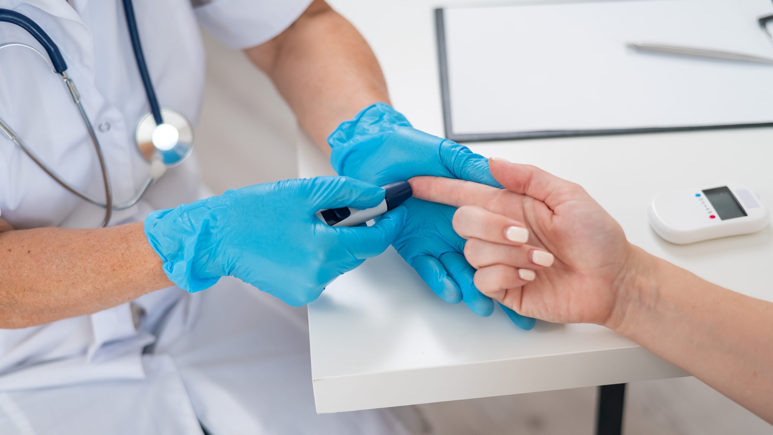 A healthcare worker wearing blue gloves uses a device to prick a patients finger for a blood test. Medical tools, a clipboard, and a glucometer are seen on the white table.
