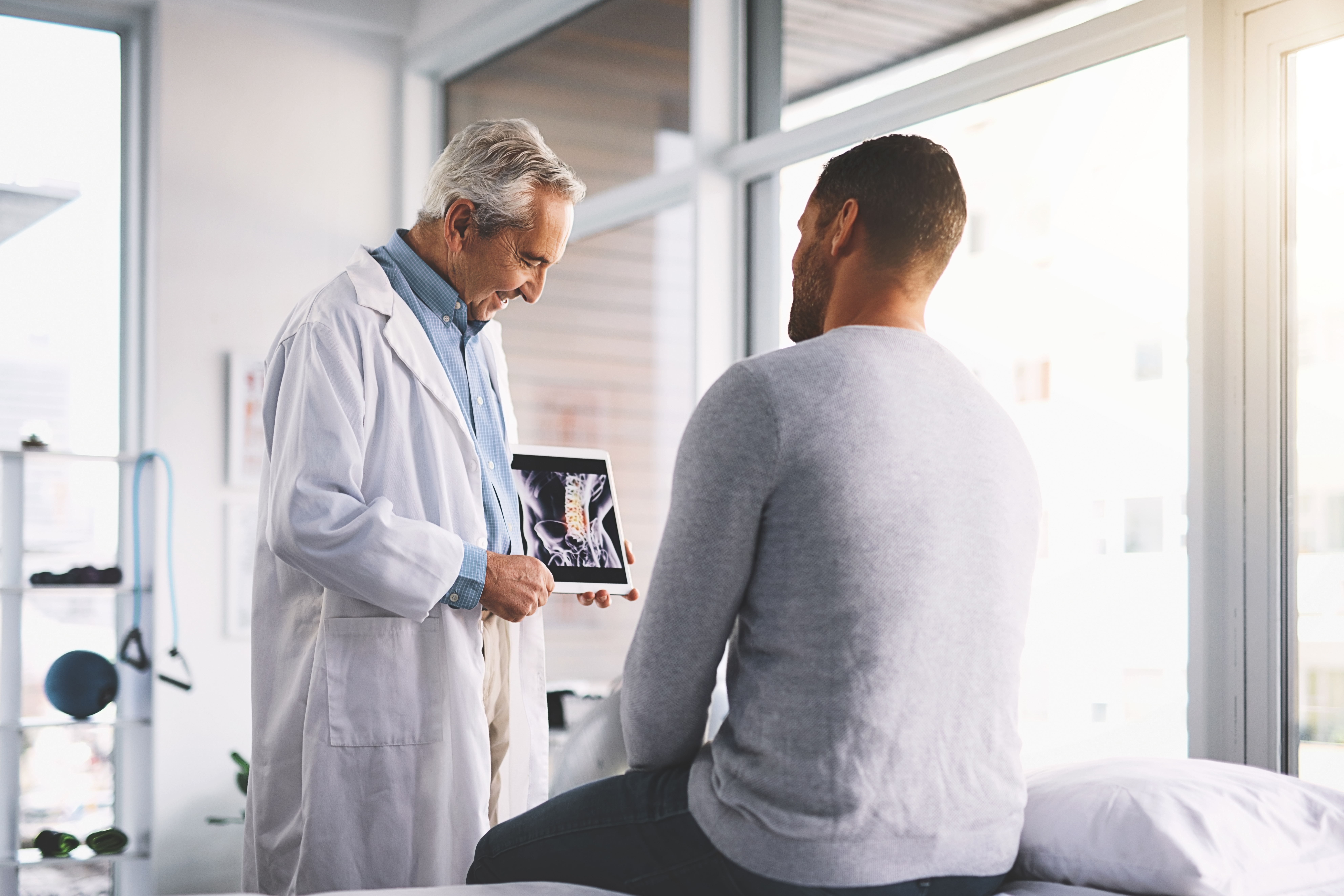 A doctor in a white coat shows a spinal X-ray on a tablet to a patient sitting on an exam table in a bright medical office with large windows.