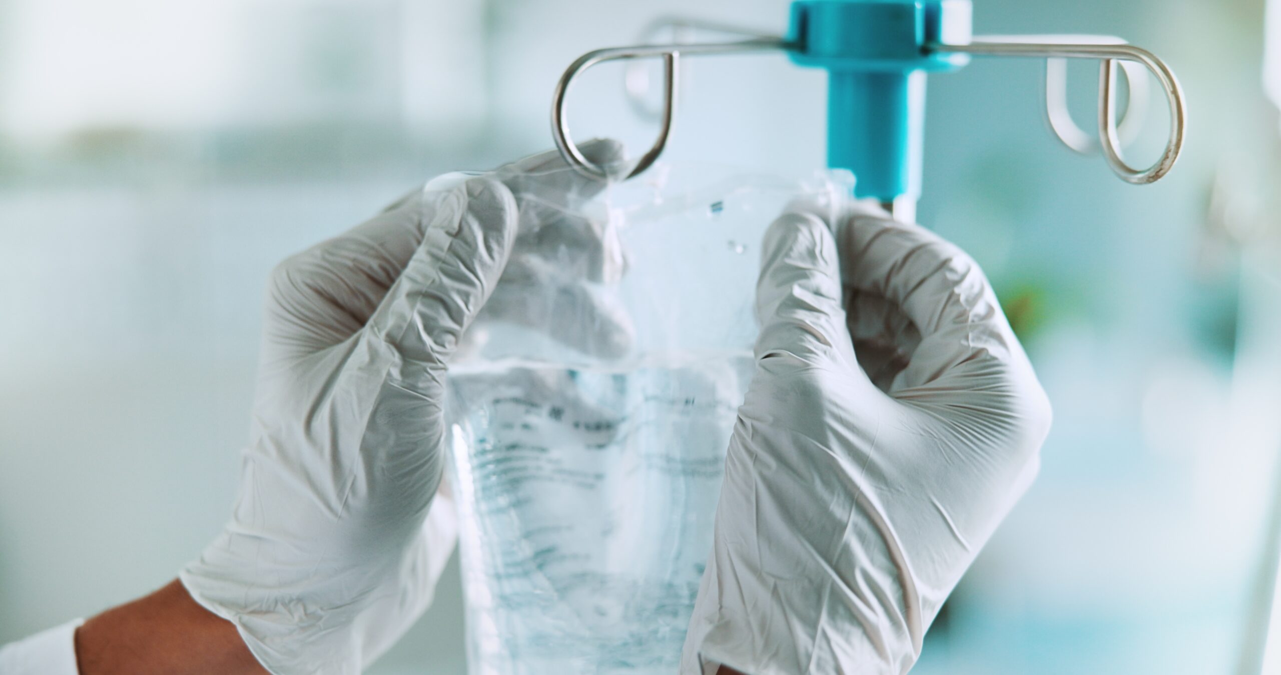 A close-up of a healthcare professional wearing white medical gloves adjusting an IV fluid bag on a hospital stand.