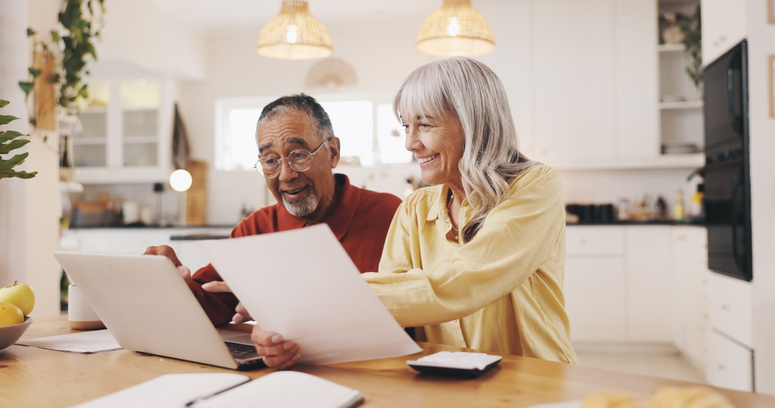 An older man and woman sit at a kitchen table, smiling while looking at papers and a laptop. The setting is bright and modern, with hanging lights and kitchen items visible in the background.