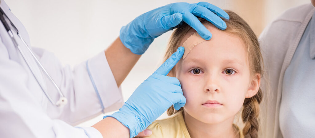 A young girl with a bandage on her forehead looks forward while a doctor in blue gloves examines her injury, gently touching her head. An adult sits beside the girl.
