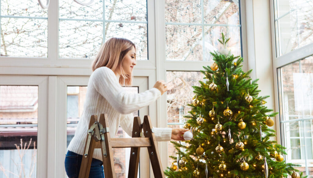 A woman decorating a Christmas tree with gold ornaments while standing on a step ladder inside a bright, modern home.