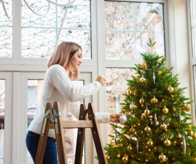 A woman decorating a Christmas tree with gold ornaments while standing on a step ladder inside a bright, modern home.