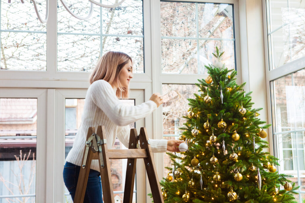 A woman decorating a Christmas tree with gold ornaments while standing on a step ladder inside a bright, modern home.