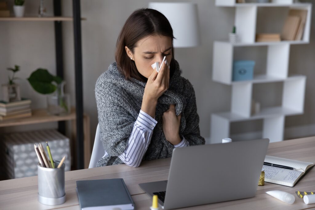 A woman sitting at a desk, wrapped in a blanket and covering her nose with a tissue, appears sick during the workday.