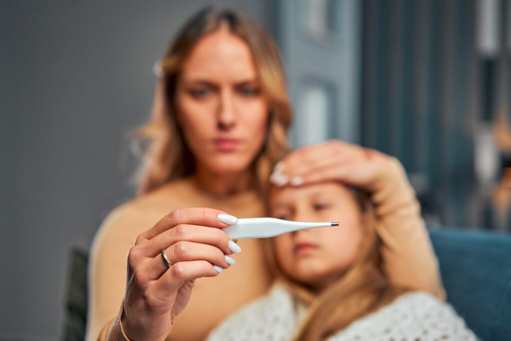 A concerned woman holds a thermometer and checks the forehead of a young girl, who is wrapped in a blanket and appears unwell.