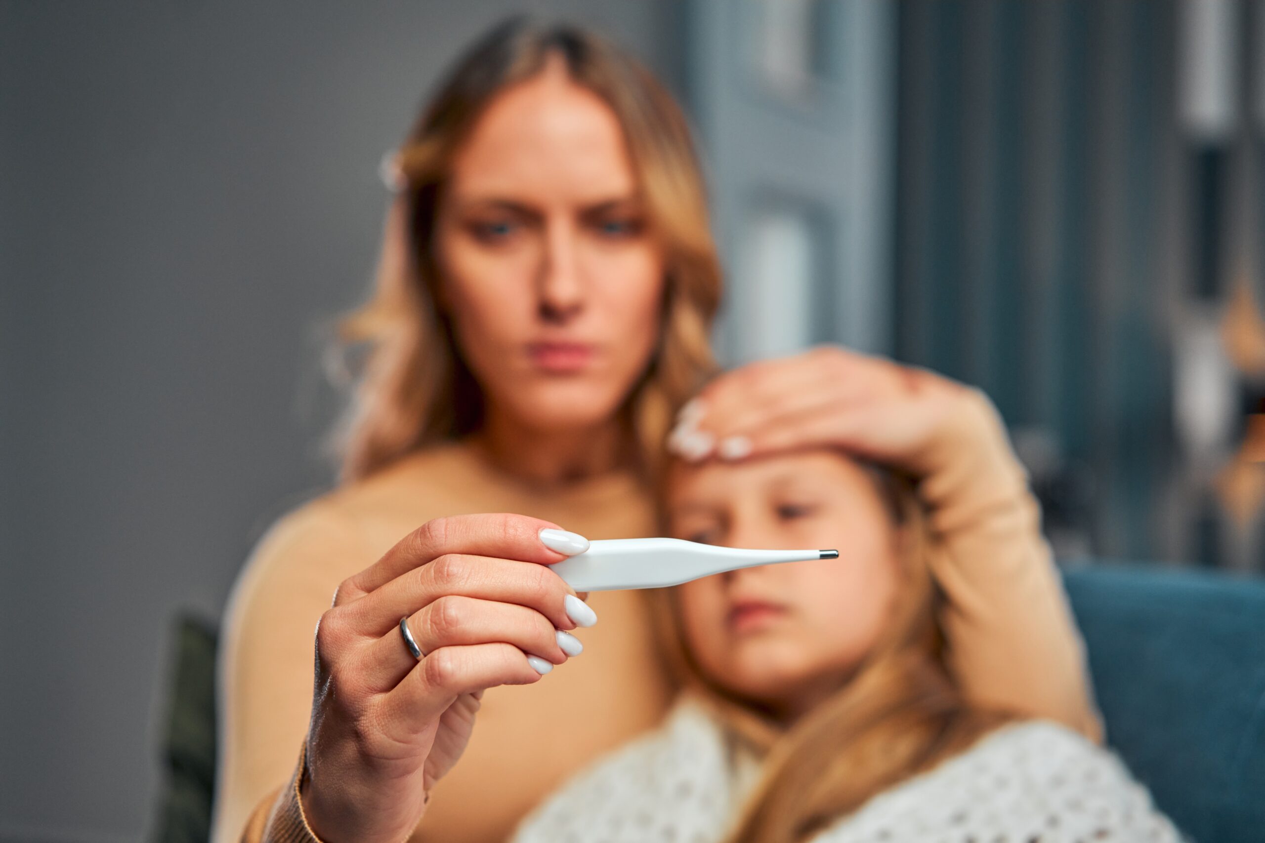A concerned woman holds a thermometer and checks the forehead of a young girl, who is wrapped in a blanket and appears unwell.