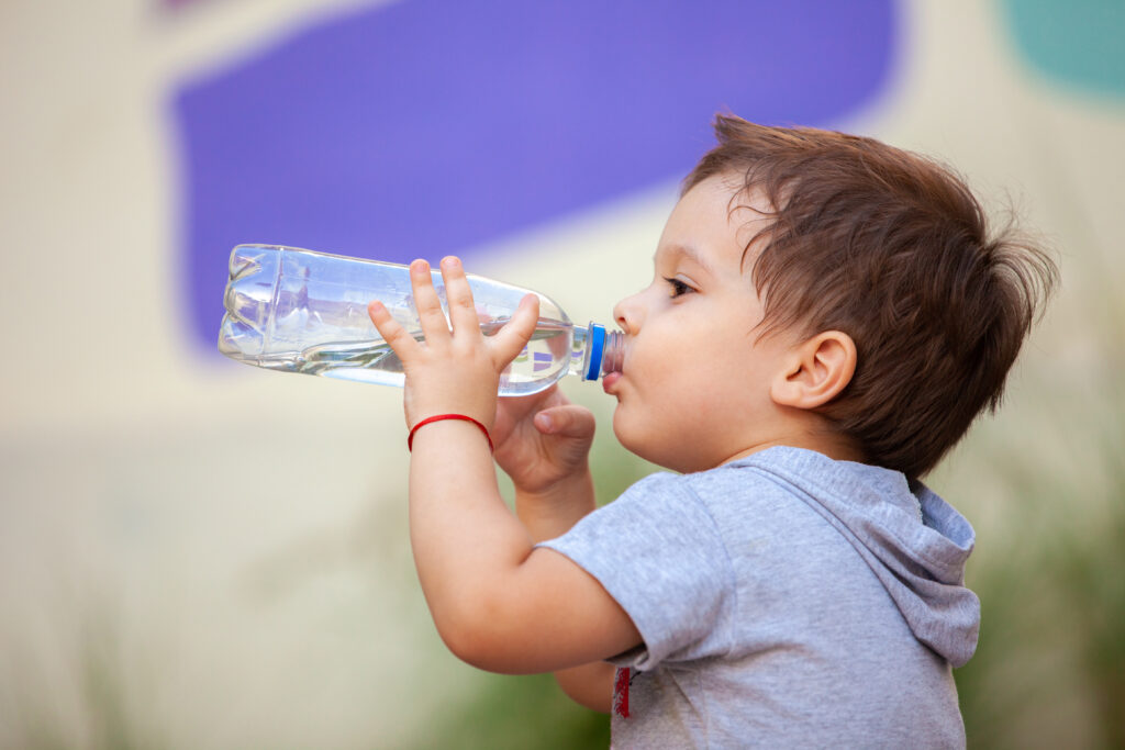 A young boy in a grey hooded t-shirt drinks water from a clear plastic bottle, holding it with both hands. He is shown in profile against a soft-focus background with a purple shapes.
