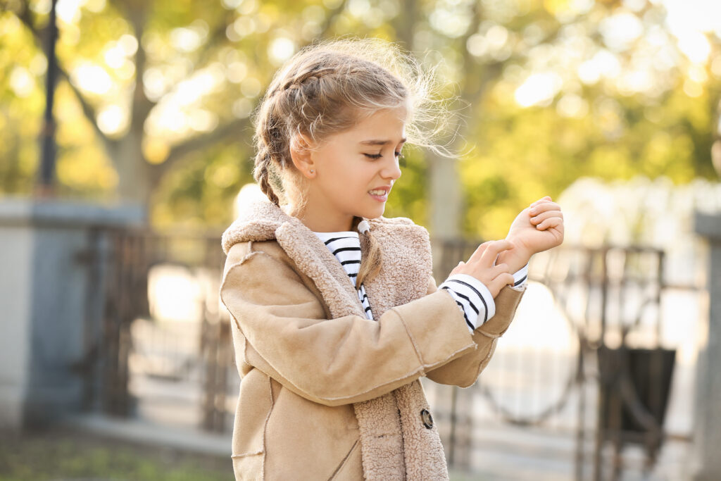 Young girl scratching her arm outdoors, appearing to experience an allergic reaction or skin irritation