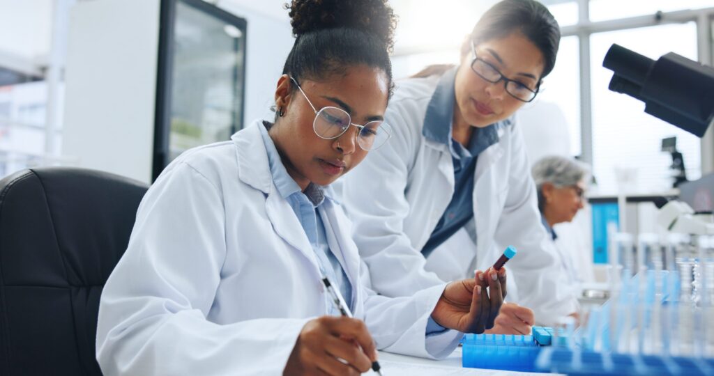 Two scientists in white lab coats examine a blood sample vial while reviewing notes in a medical laboratory.