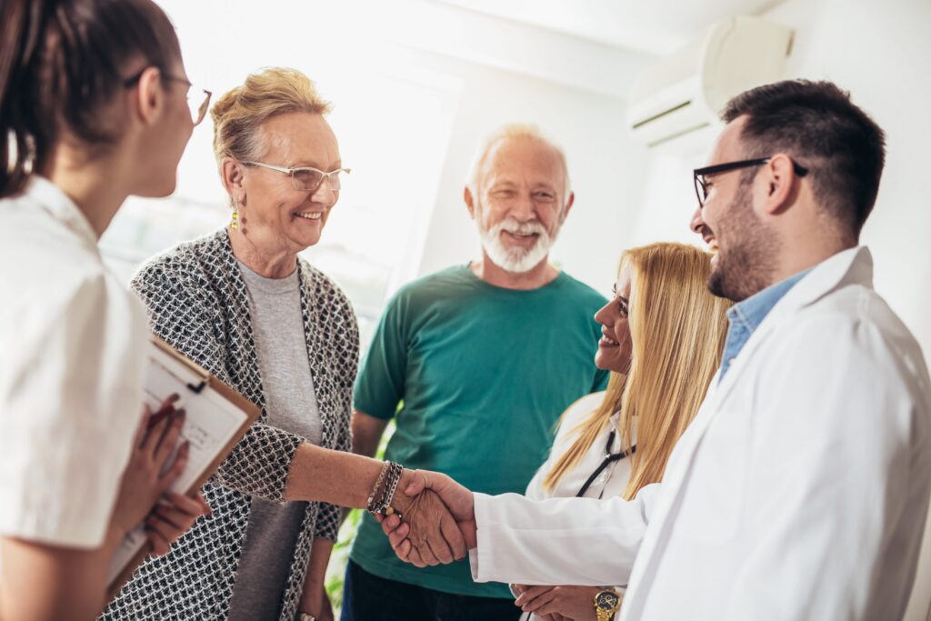 Three healthcare professionals in white coats shaking hands with elderly couple in a bright room.