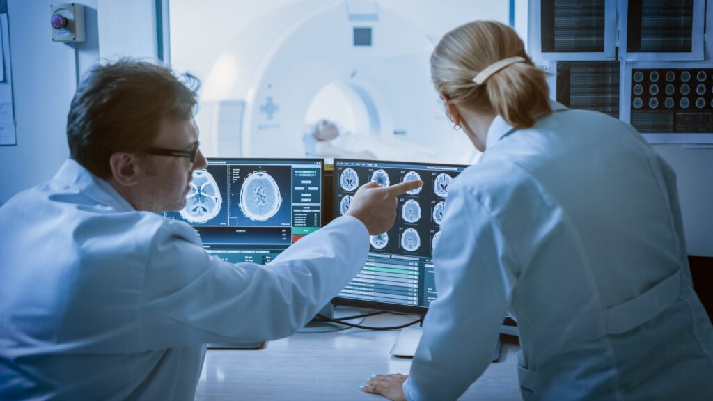 Doctor and radiologist in control room reviewing brain scan monitors while patient undergoes MRI or CT scan procedure in background