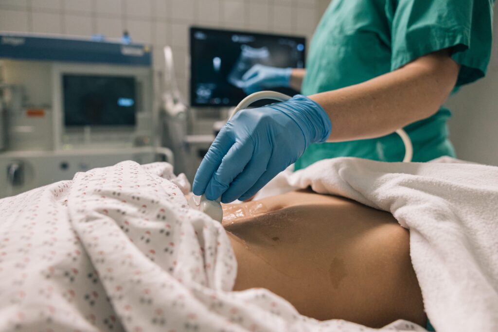 Doctor in scrubs and gloves performing abdominal ultrasound examination on patient lying on hospital bed.