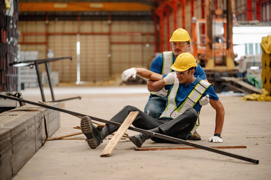 Construction worker lying on ground receiving aid from team members after metal bars and wood planks fall on him at worksite.