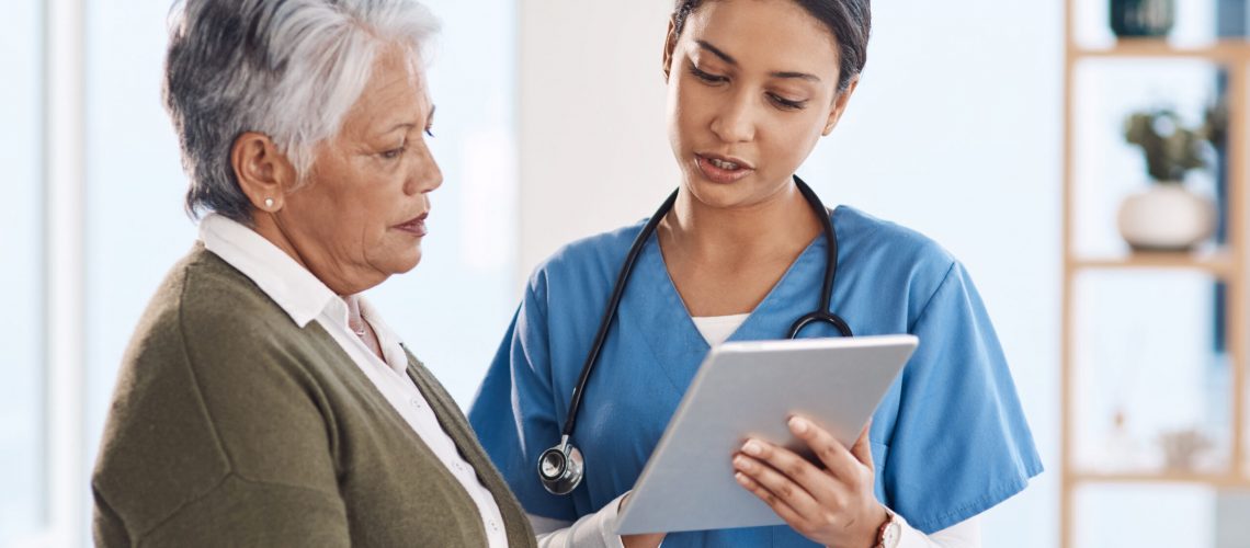 A young female doctor in blue scrubs and a stethoscope is showing information on a tablet to an older female patient with gray hair.