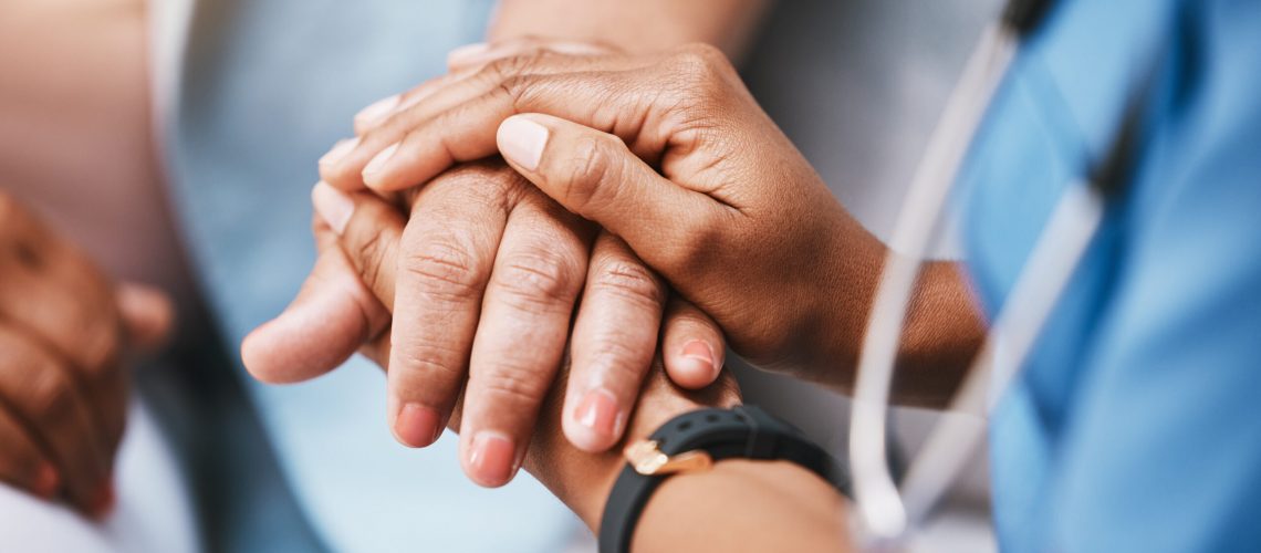 A close-up of a doctor or nurse in blue scrubs gently holding the hands of an elderly patient to offer comfort and support.