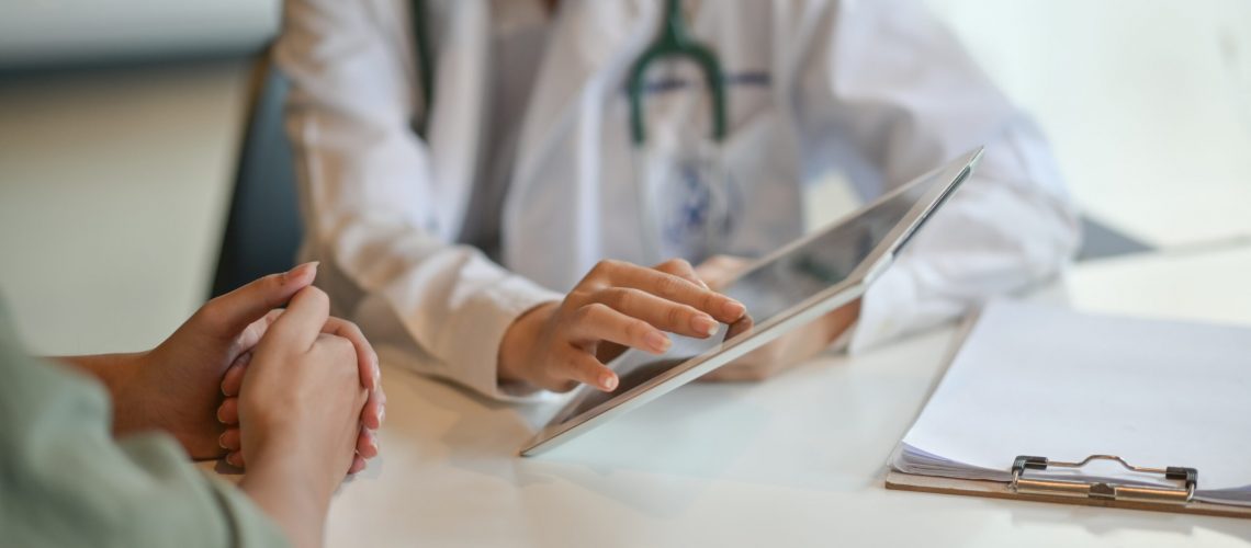 A doctor in a white coat, holding a tablet, discusses information with a patient. Both are seated at a desk, with a clipboard and papers nearby, suggesting a medical consultation.